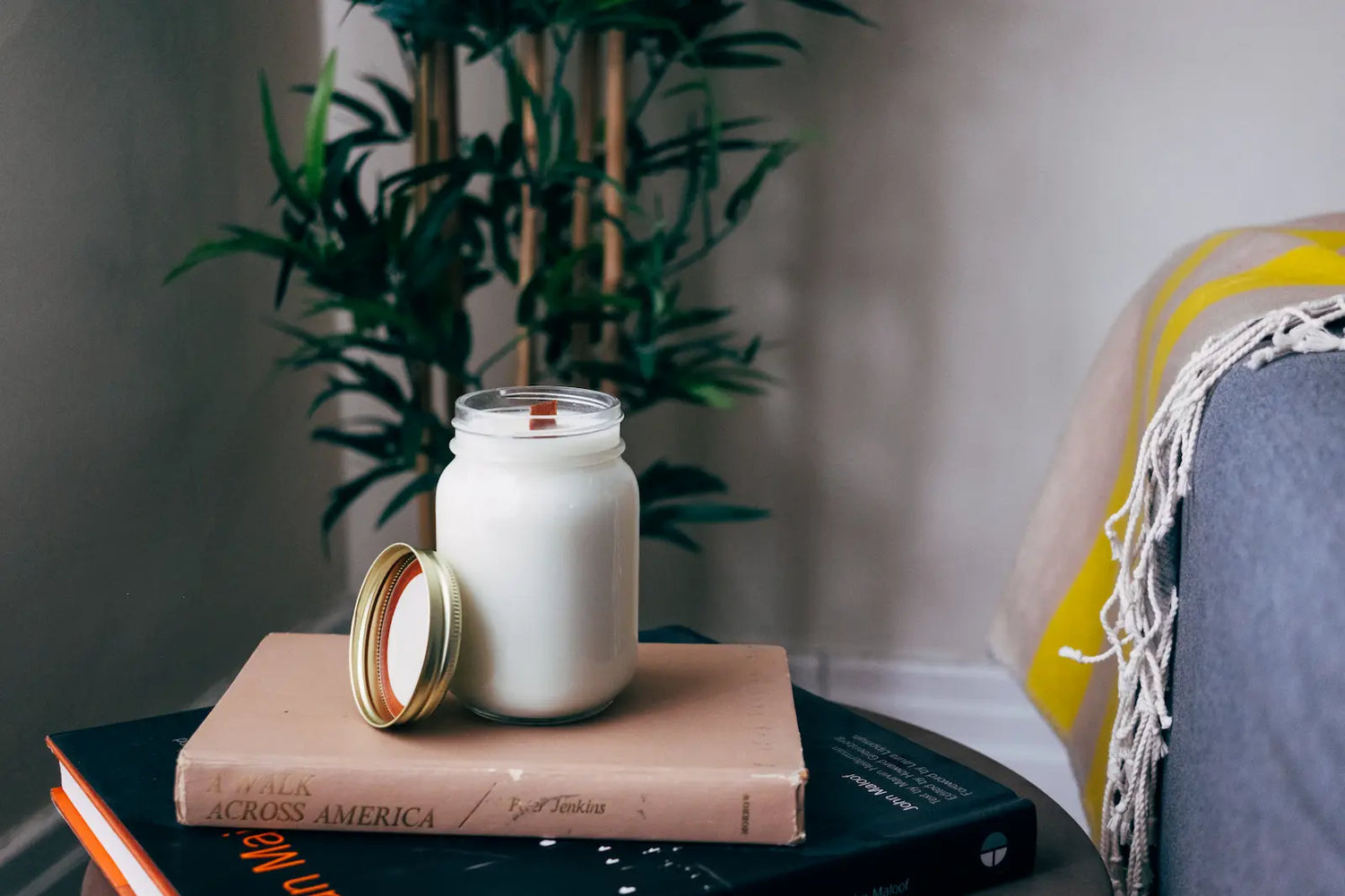 Organic handmade candle with a wooden wick in a glass mason jar, placed on stacked books with a green plant in the background.